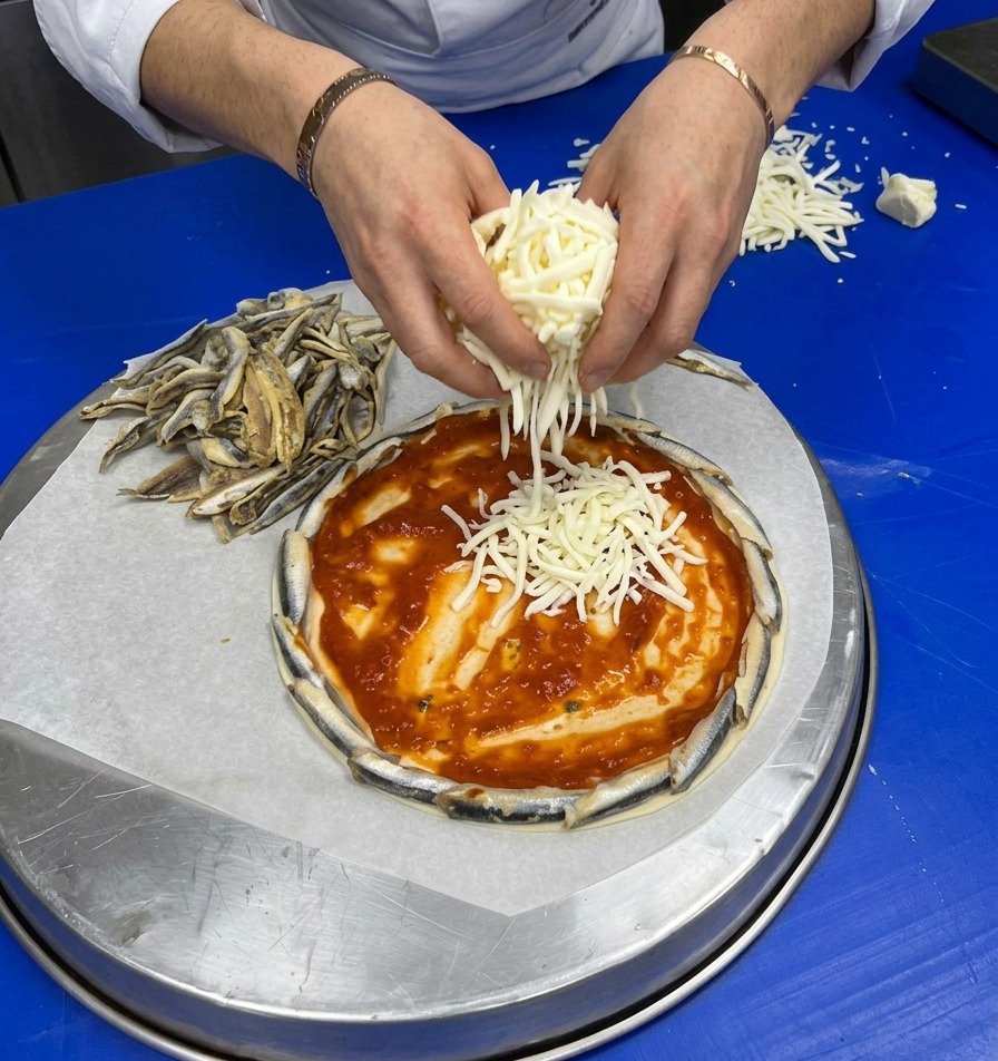 Dłonie hojnie posypujące świeżo startym, białym serem spód pizzy posmarowany czerwonym sosem i z brzegiem z sardeli.