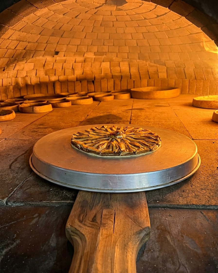 Pizza z sardelami leżąca na okrągłej metalowej tacy piecze się wewnątrz rozżarzonej, pomarańczowej komory murowanego pieca opalanego drewnem.