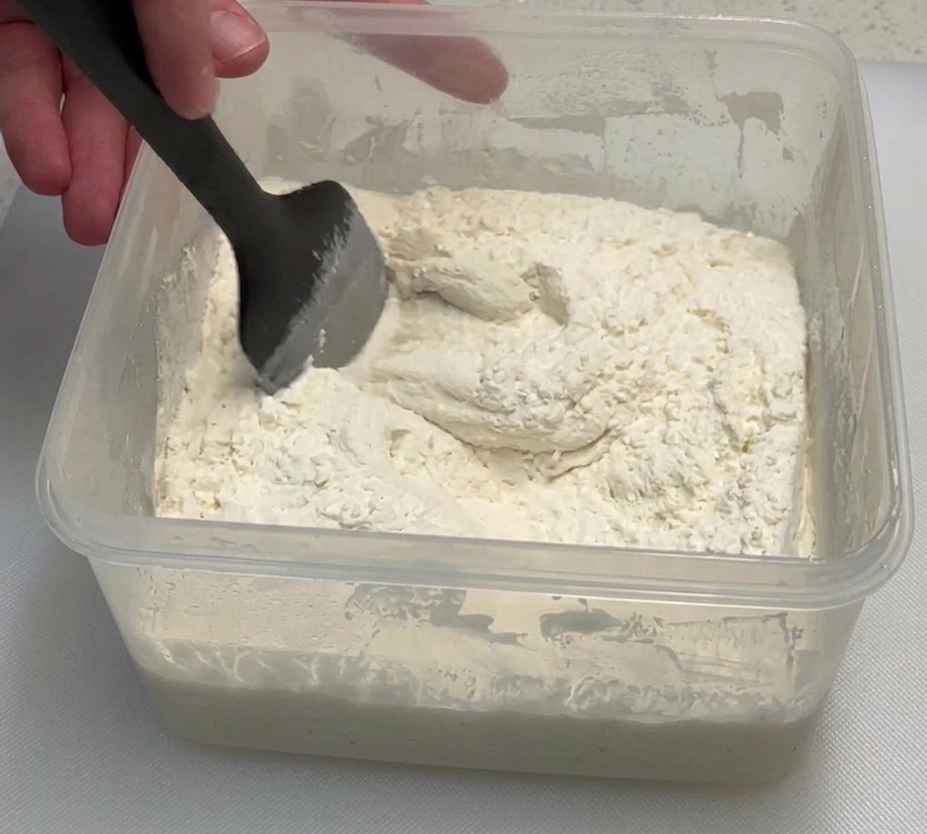 Dłoń z czarną szpatułką mieszająca lepką mąkę z zaczynem poolish w przezroczystym pojemniku.