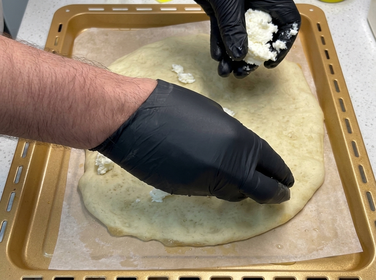 Dłonie w czarnych rękawiczkach układające porwane kawałki białej mozzarelli na złotym, podpieczonym spodzie pizzy.