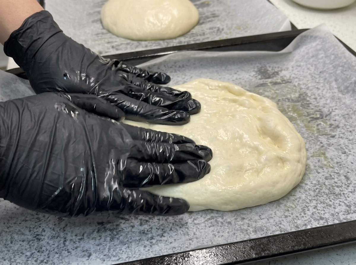 Dłonie w rękawiczkach delikatnie rozciągające ciasto na pizzę na blasze z papierem do pieczenia.