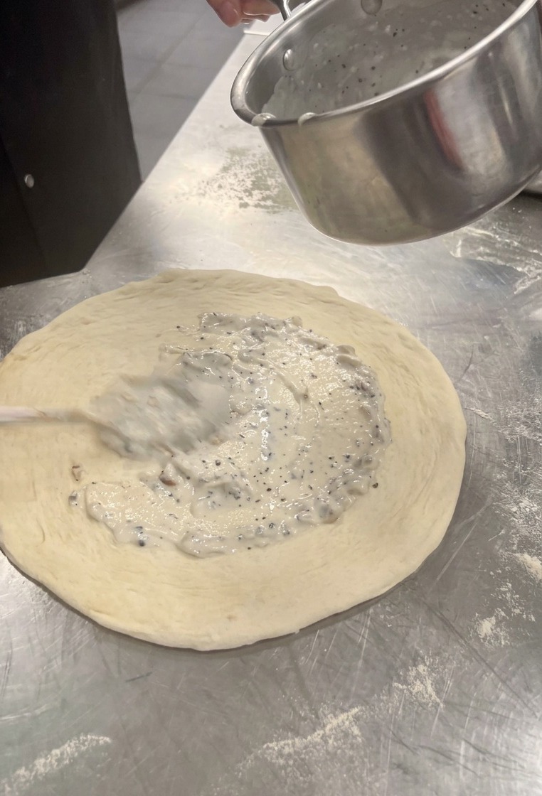 Nakładanie łyżką białego, kremowego sosu grzybowego na przygotowany spód do pizzy.
