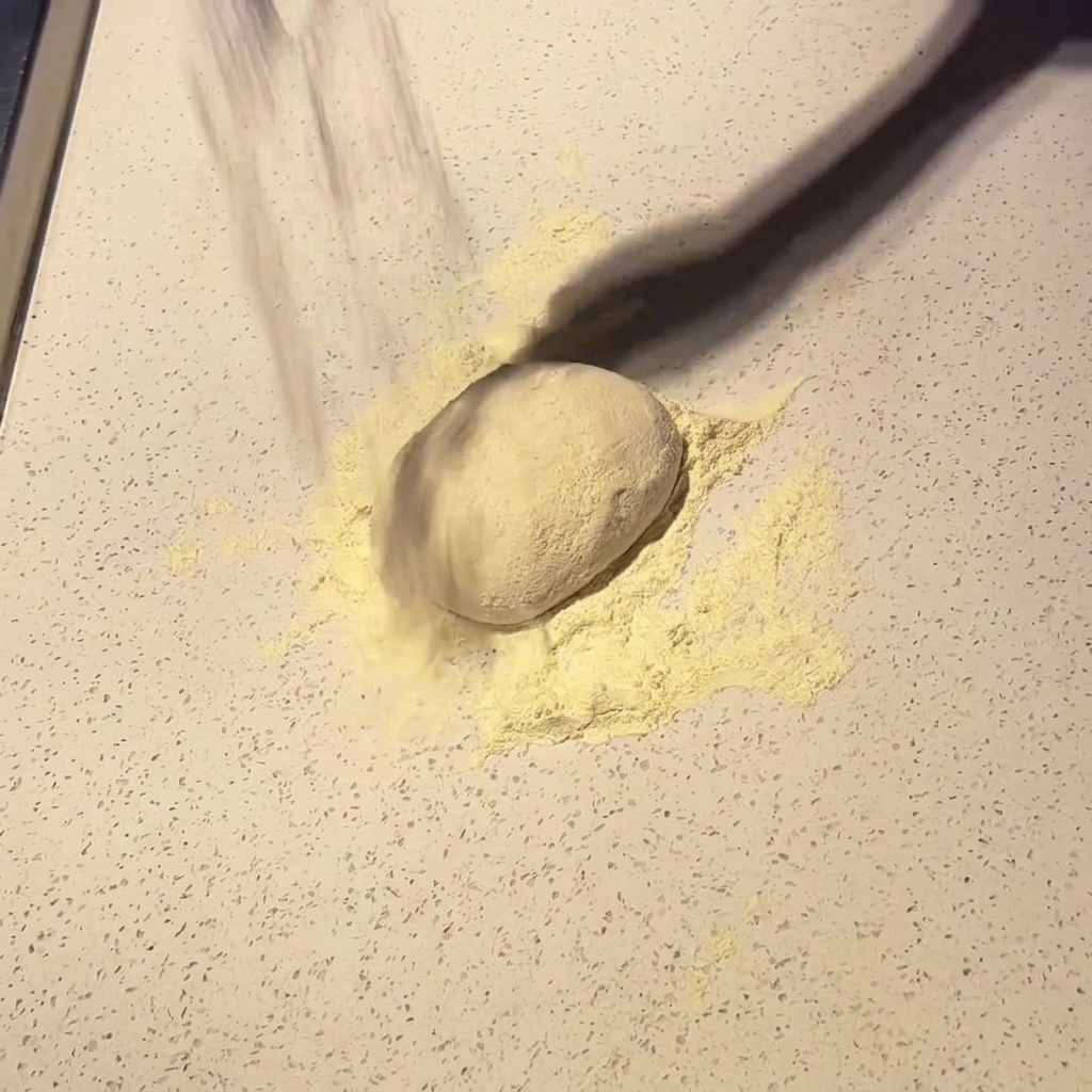 Kula ciasta na pizzę posypana mąką na jasnym blacie.