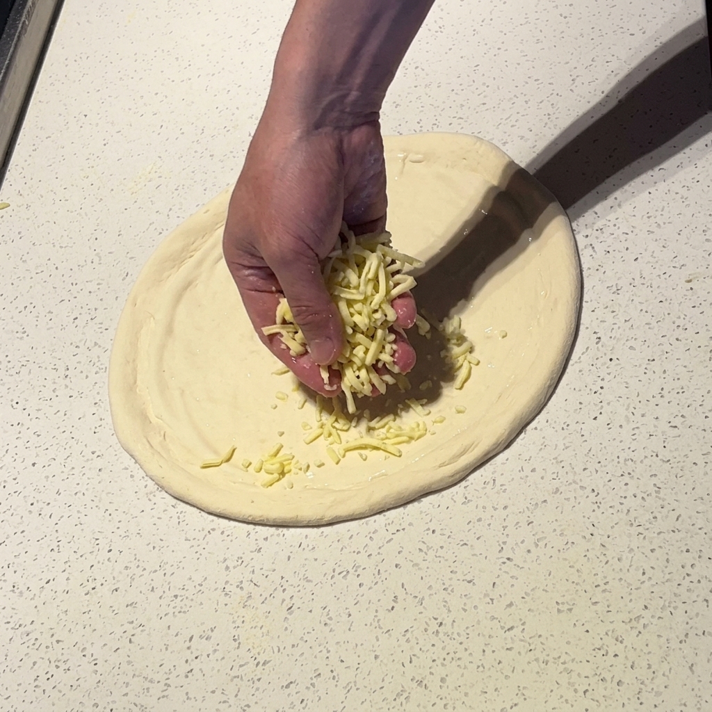Dłoń posypująca tartą mozzarellą rozciągnięte ciasto na pizzę pokryte białym sosem.