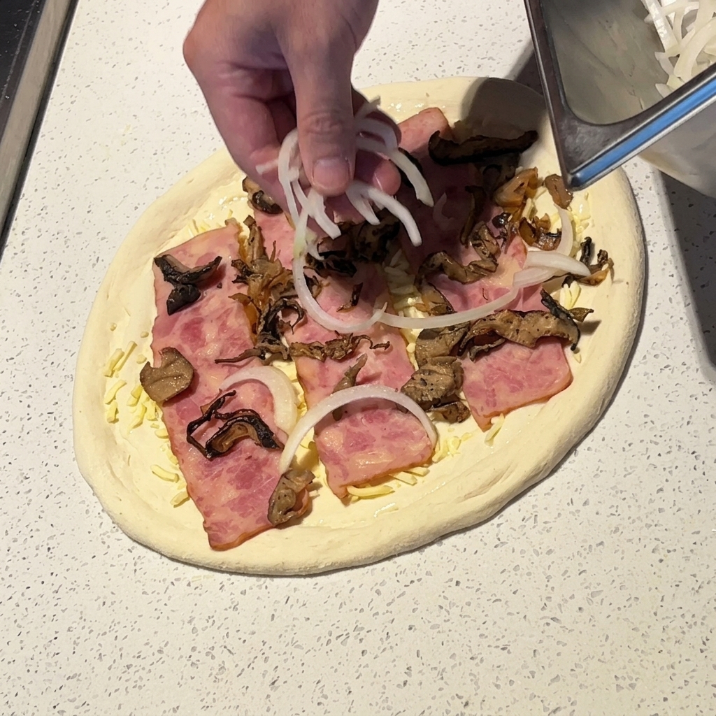 Dłoń rozrzucająca cienkie plastry białej cebuli na pizzy z boczkiem i grzybami.