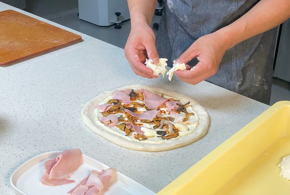 Dłonie kucharza rwące świeżą mozzarellę nad pizzą z szynką i grzybami.