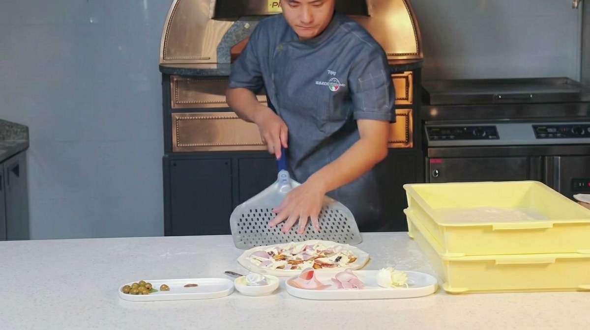 Kucharz wciera mąkę w perforowaną metalową łopatę do pizzy.