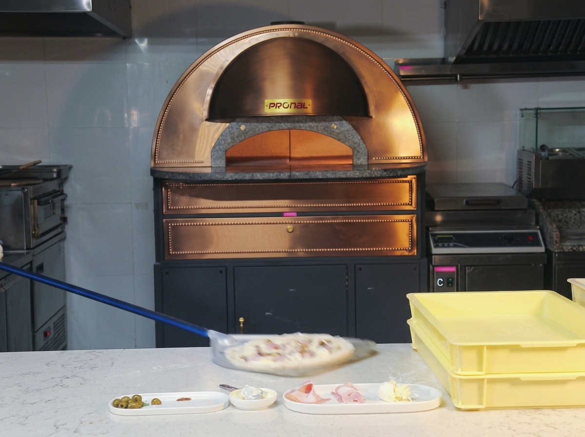 Surowa pizza z szynką i grzybami na długiej metalowej łopacie wsuwana do rozgrzanego pieca kopułowego.