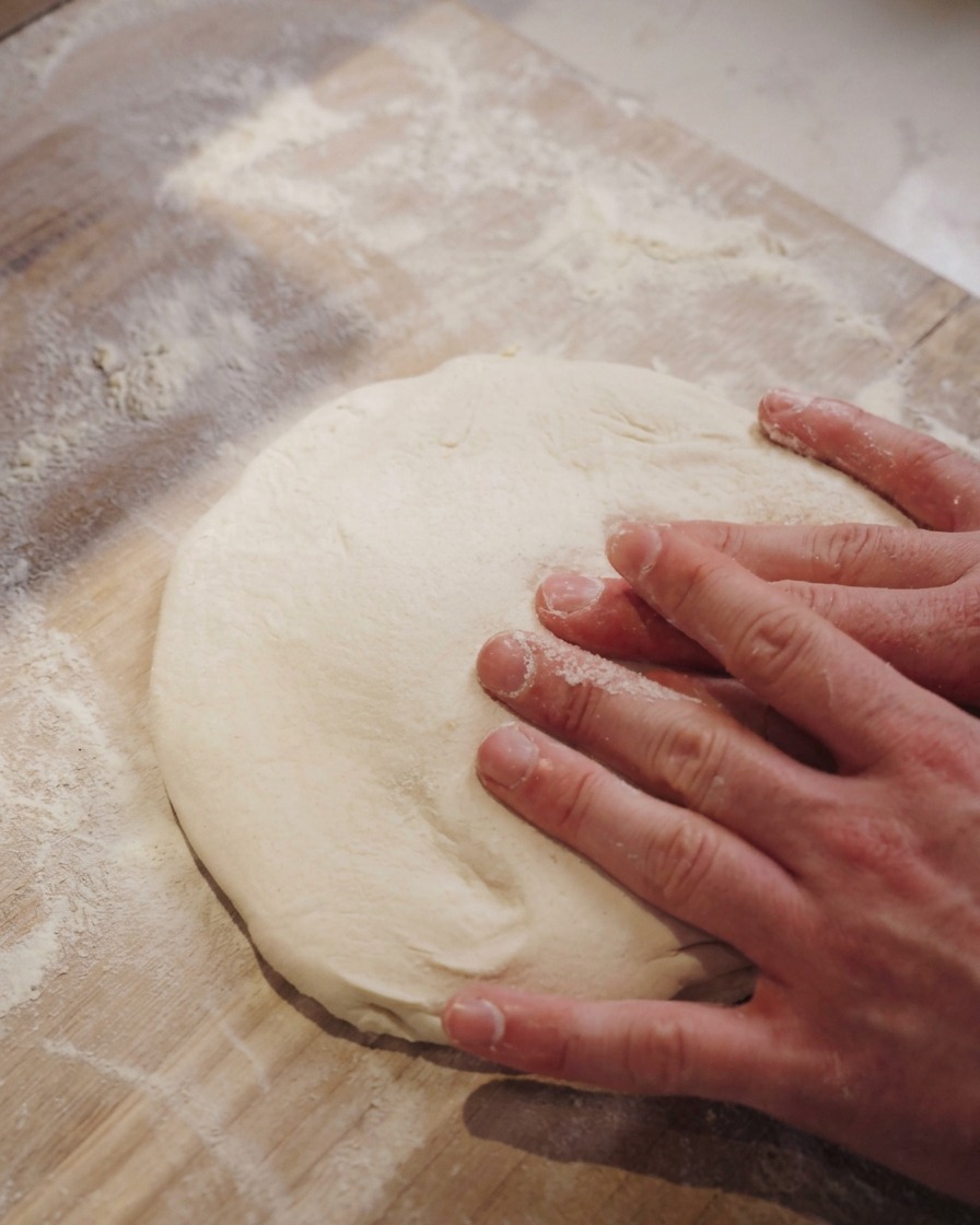 Dłonie delikatnie naciskające środek miękkiego ciasta na pizzę na blacie posypanym mąką.
