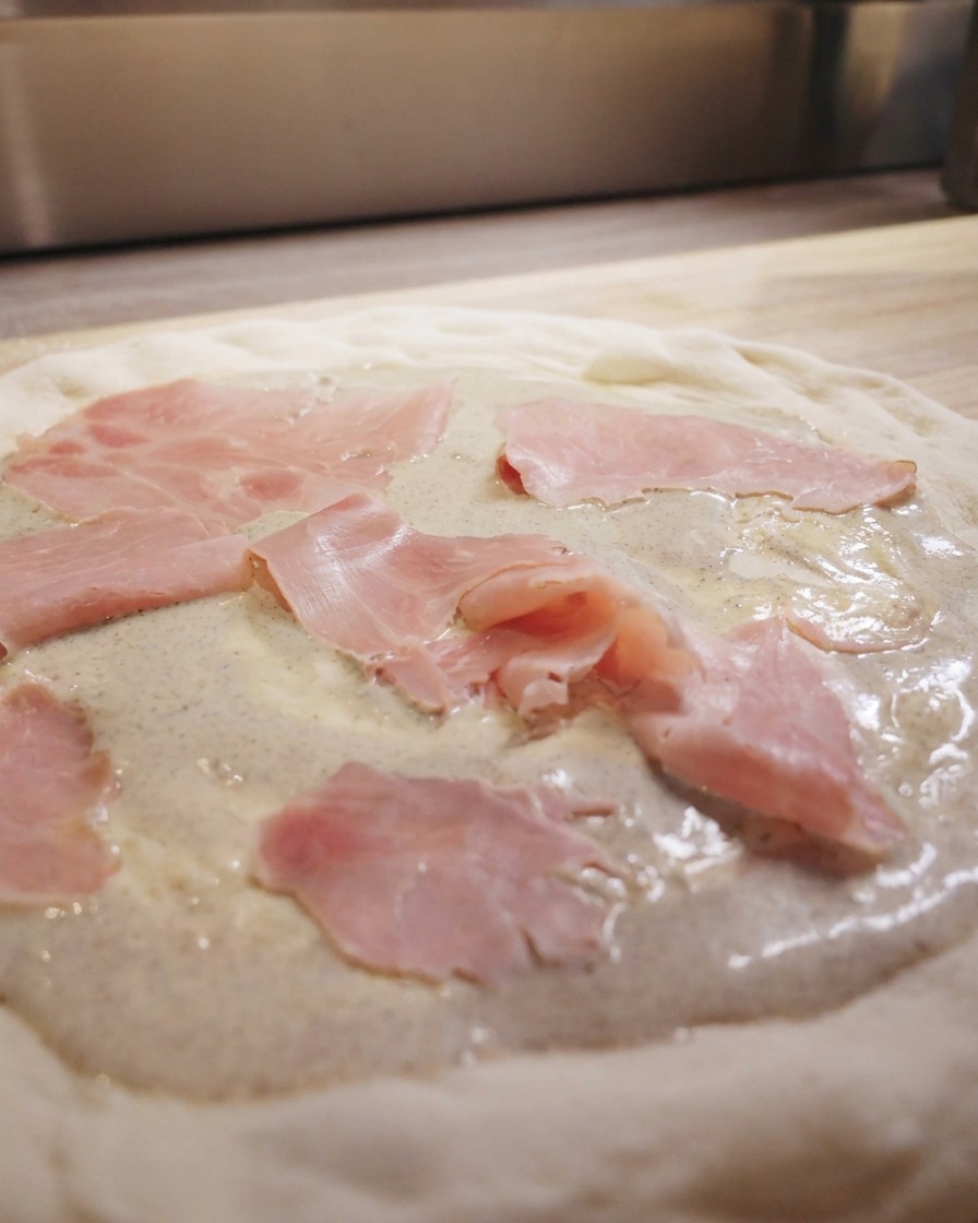 Plastry różowej szynki ułożone na spodzie do pizzy z kremem truflowym.