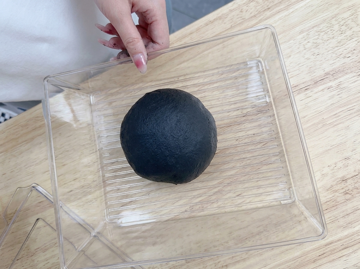 Dłoń wkładająca okrągłą, czarną kulę ciasta do przezroczystego, plastikowego pojemnika na drewnianym blacie.