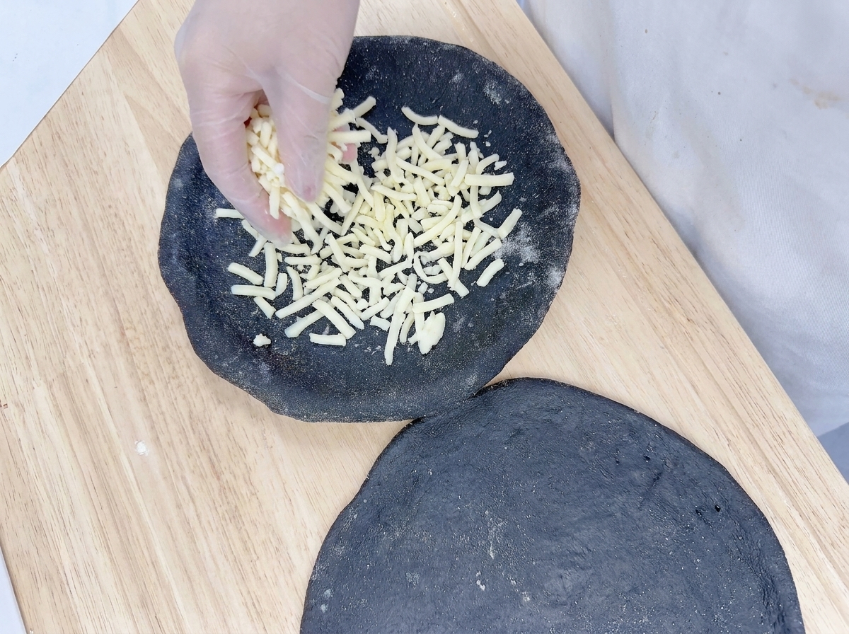 Dłoń równomiernie rozsypująca tartą mozzarellę na czarnym spodzie do pizzy.
