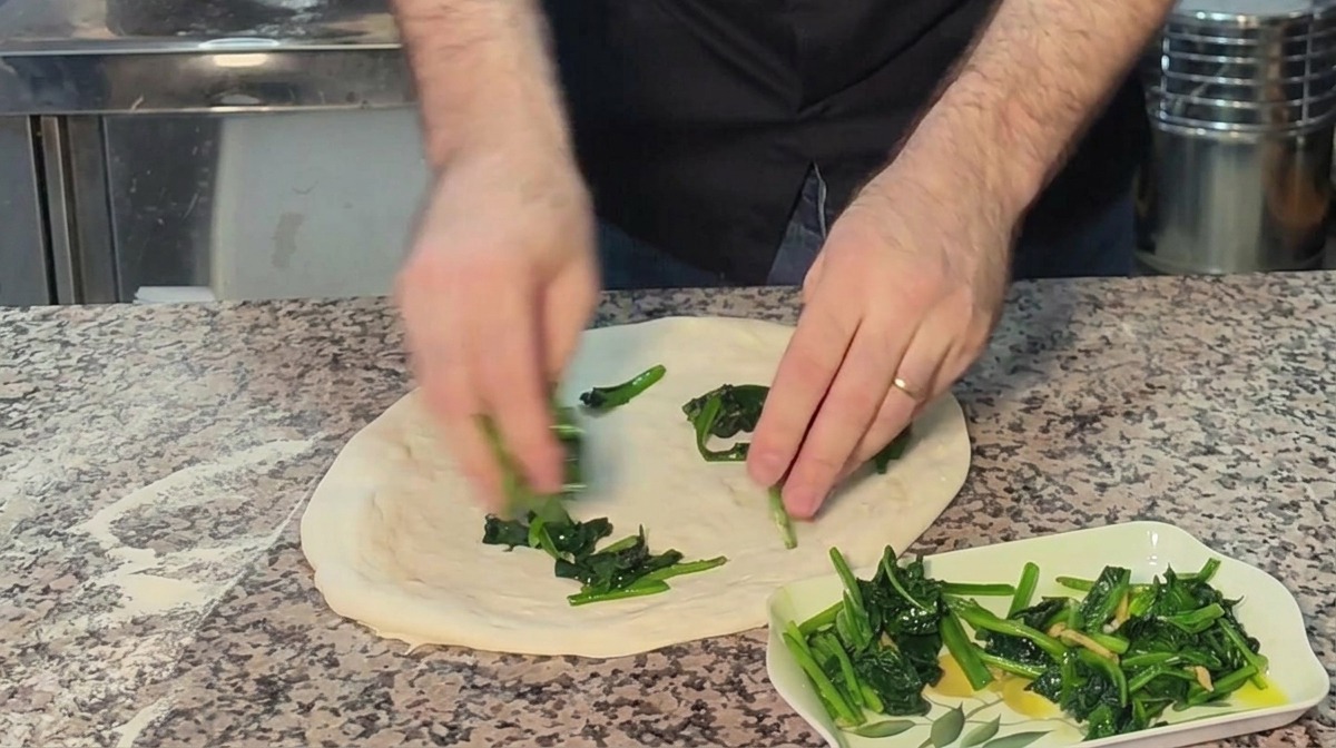 Dłonie rozkładające zielony szpinak na surowym spodzie pizzy.
