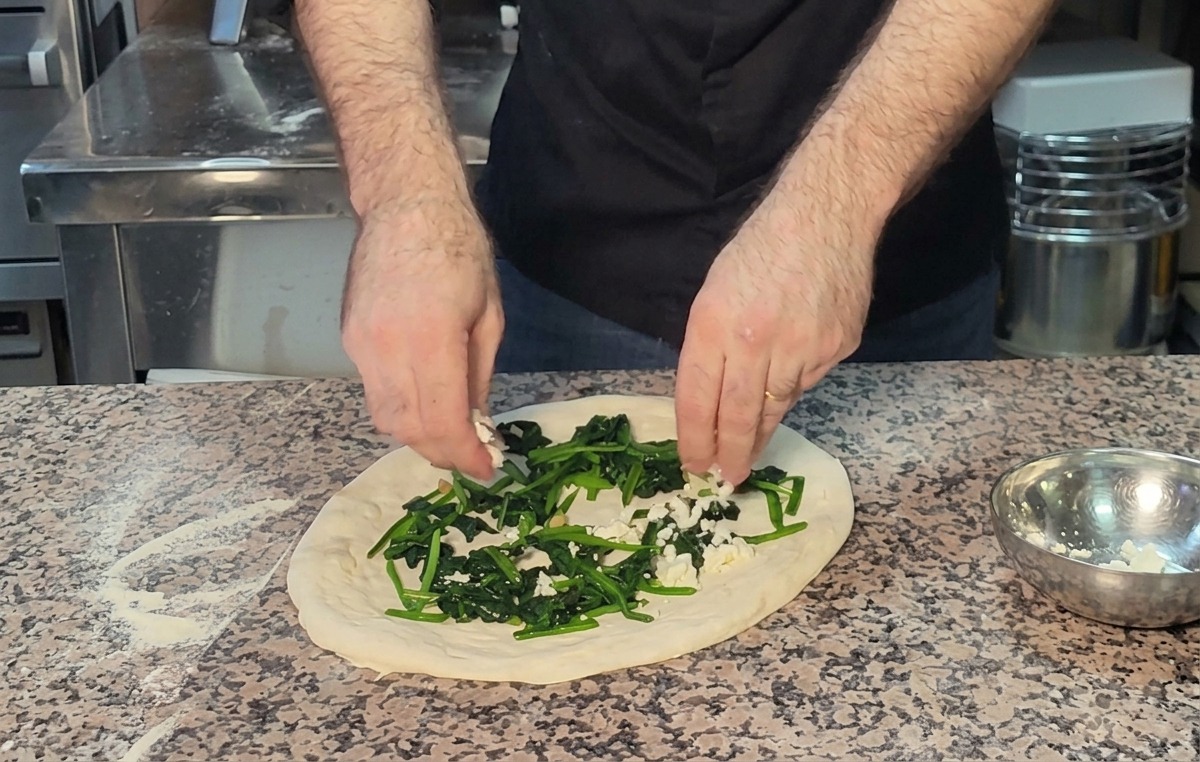 Dłonie posypujące pokruszoną fetą warstwę szpinaku na pizzy.