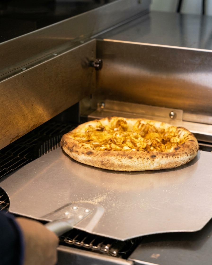 Wyjmowanie upieczonej pizzy z pieca za pomocą metalowej łopaty; brzegi są lekko przypieczone.