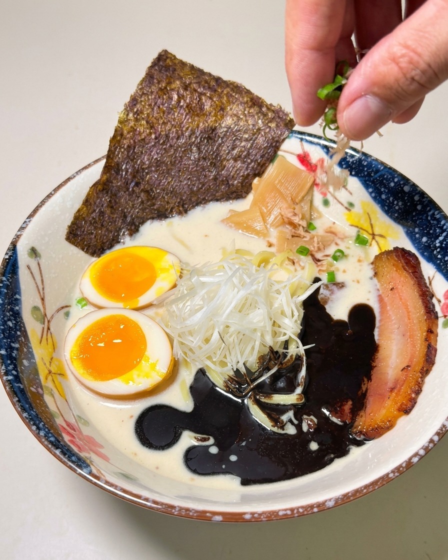 Dłoń posypująca posiekaną dymką pięknie podany ramen z czarnym olejem.