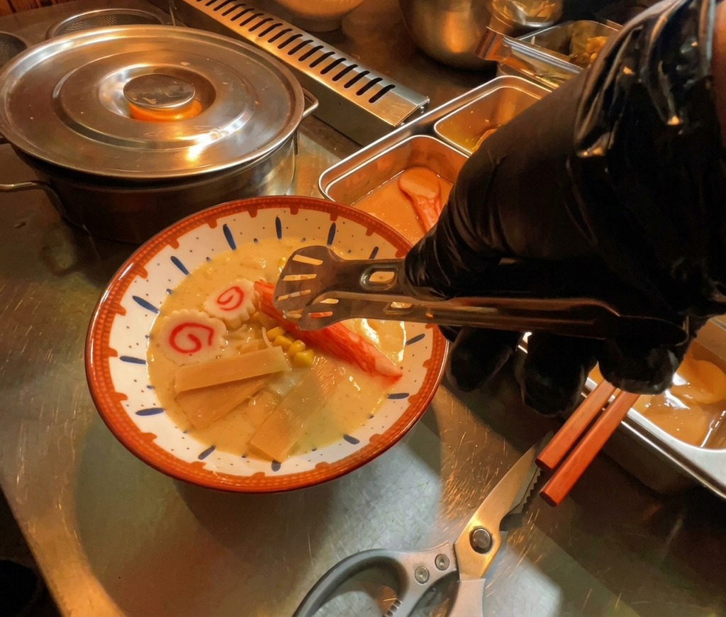 Dłoń w rękawiczce kładzie plastry ciastka rybnego narutomaki do parującej miski ramen.