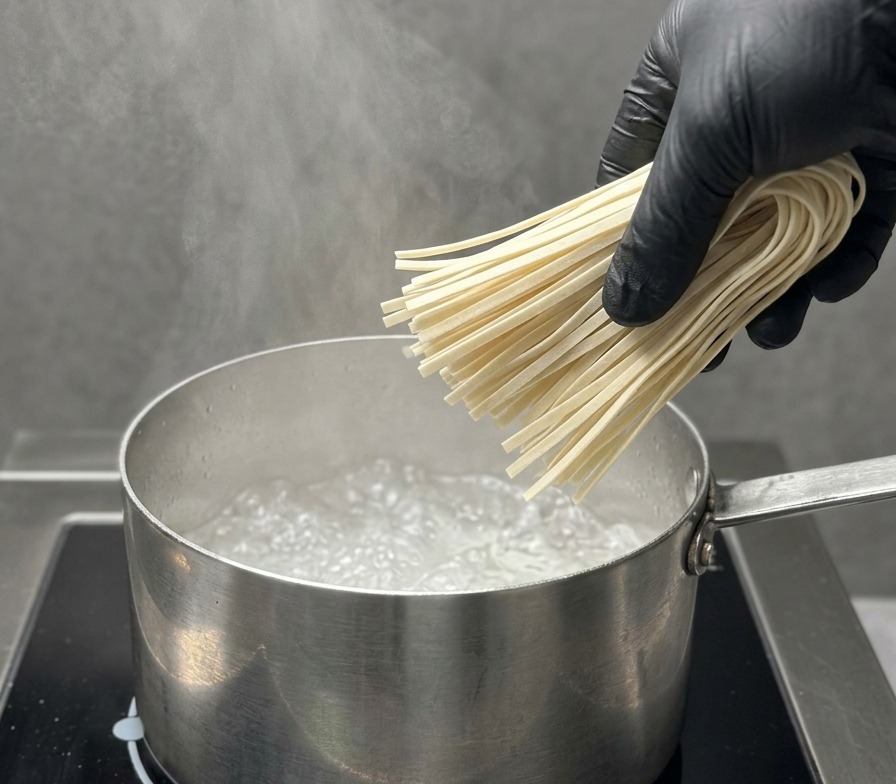 Ręka w czarnej rękawiczce ostrożnie wkłada porcję prostego, suszonego makaronu ramen do garnka z wrzącą wodą.