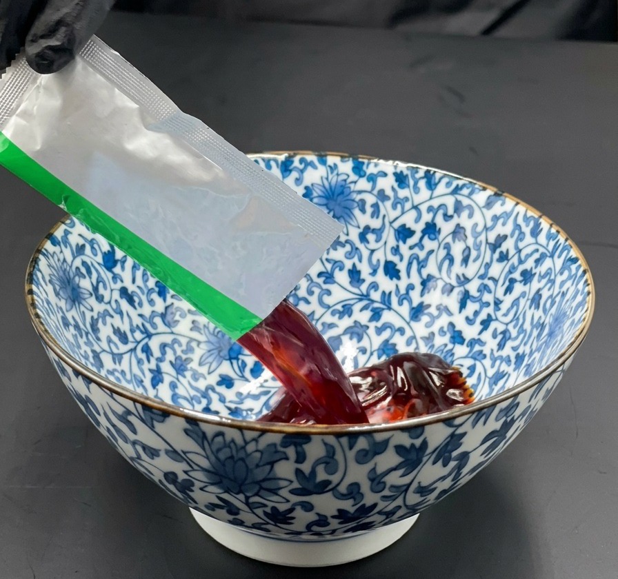Ręka wlewająca ciemną bazę do ramenu z saszetki do ceramicznej miski w niebiesko-białe wzory.