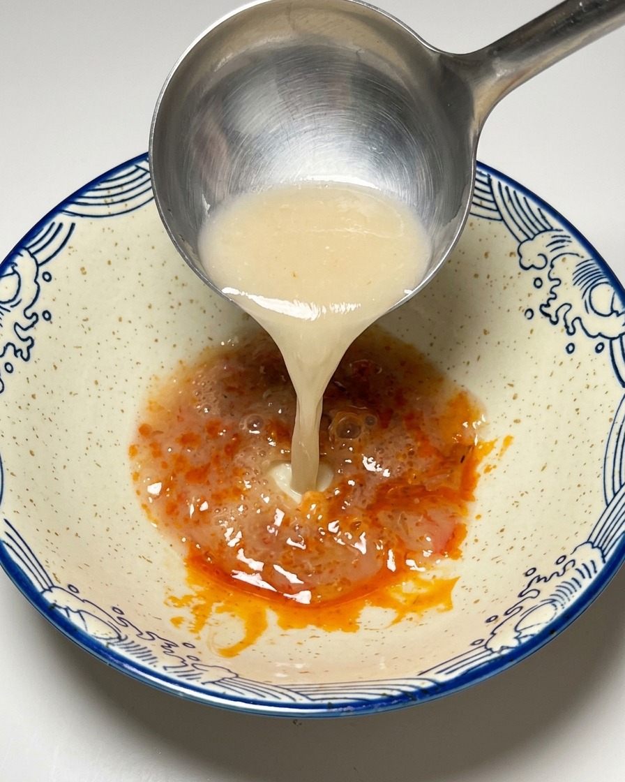 Kremowy wywar tonkotsu nalewany z chochelki do miski z pastą chili.