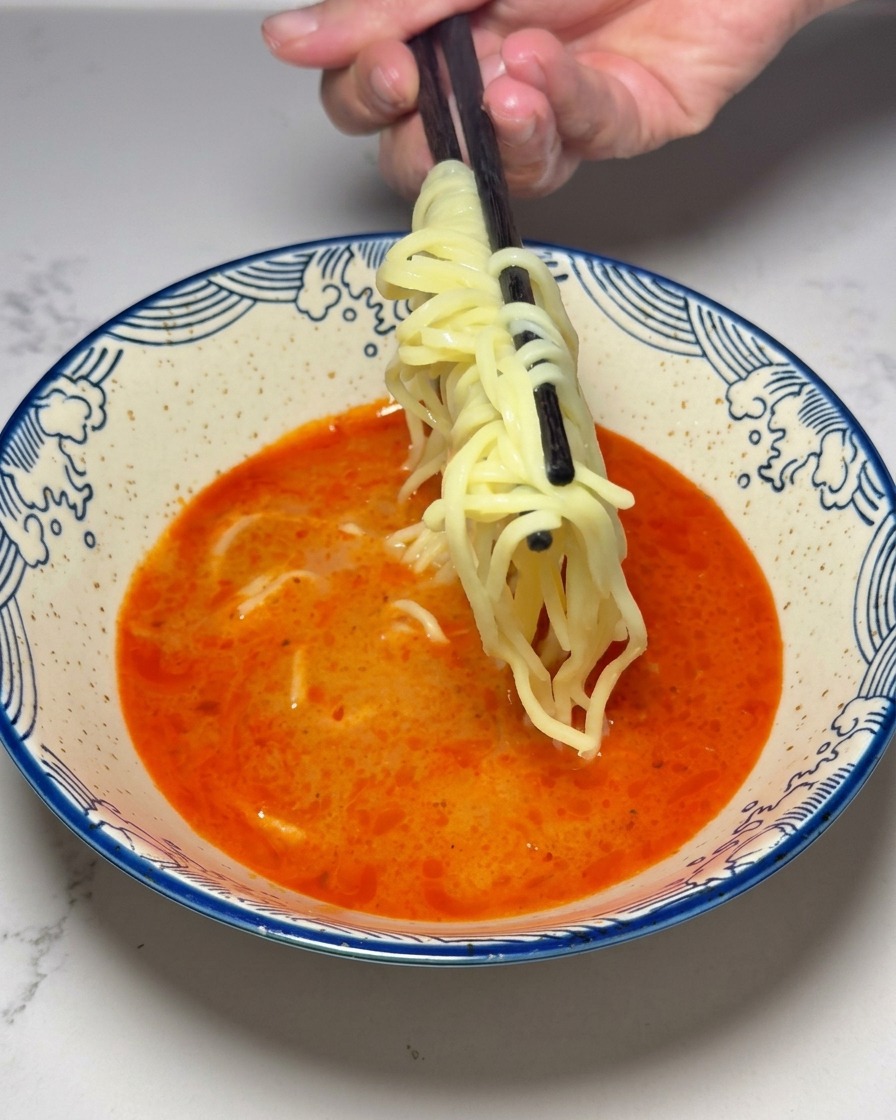 Ugotowany żółty makaron ramen wkładany do miski z czerwoną zupą.