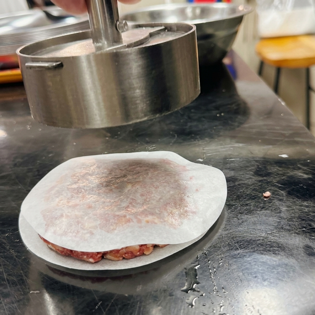 Metalowa prasa do burgerów formująca mieloną wołowinę w idealny krążek na kawałku papieru do pieczenia.