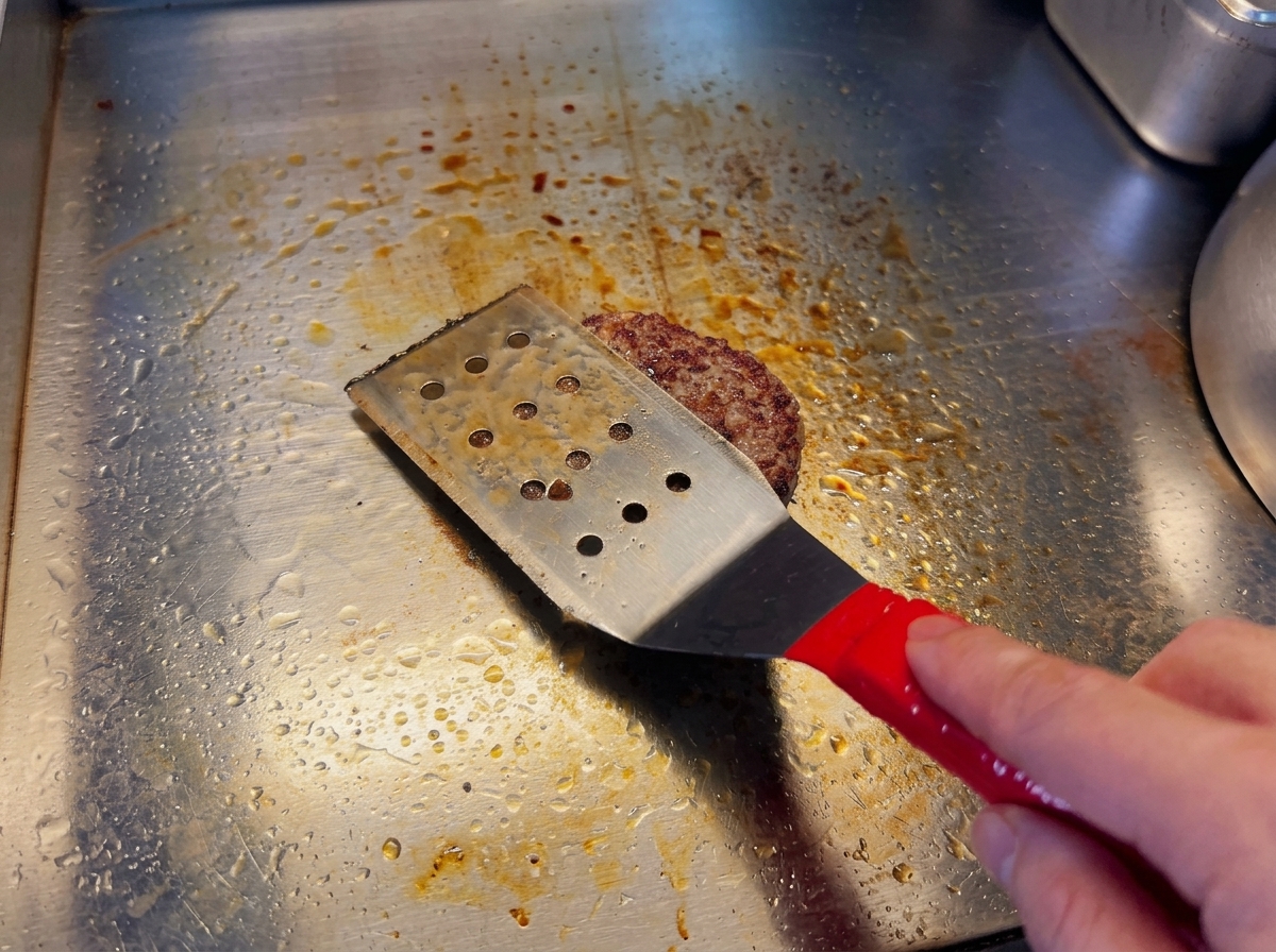 Dłoń trzymająca łopatkę, delikatnie dociskająca smażący się kotlet wołowy do gorącej powierzchni.