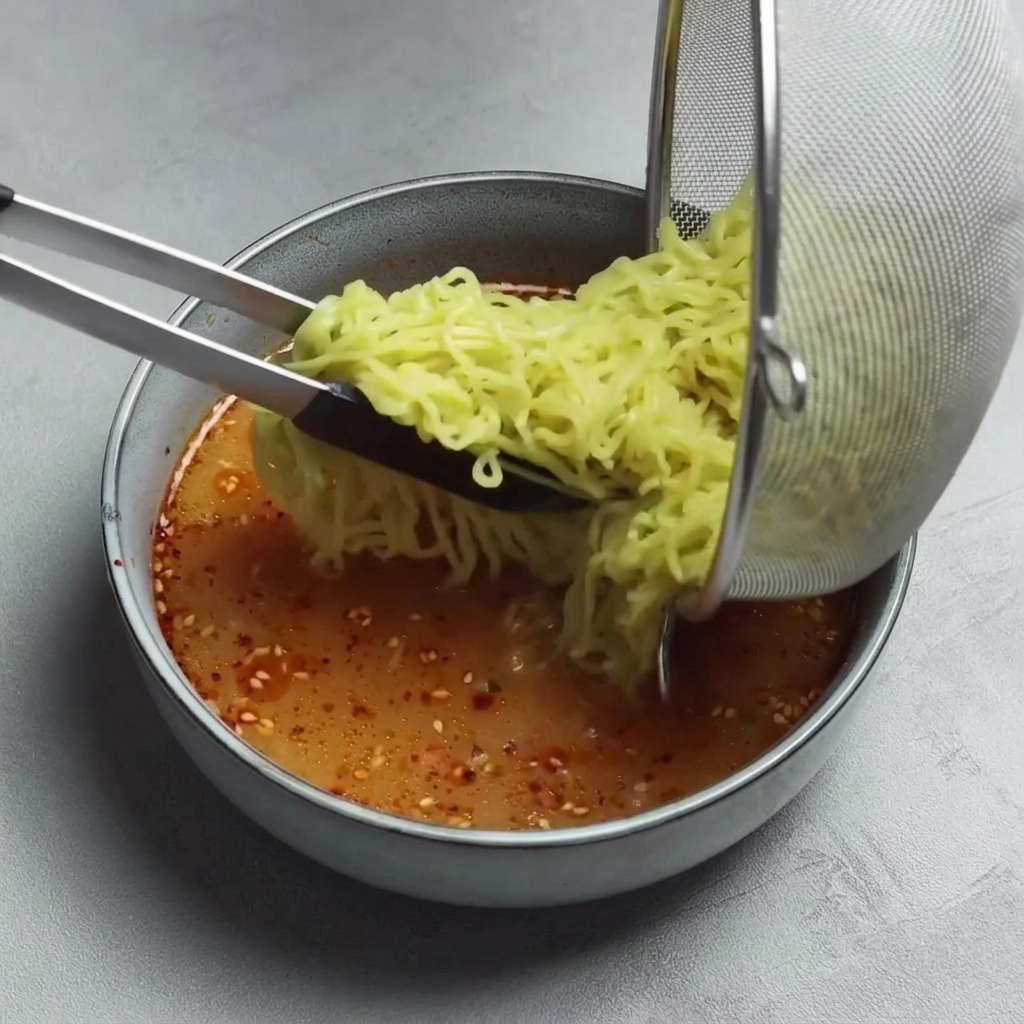 Szczypce przekładające ugotowany makaron ramen z metalowego sitka do miski z czerwonym, pikantnym bulionem.