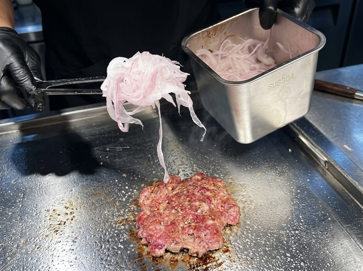 Garść bardzo cienko pokrojonej czerwonej cebuli kładziona na smażący się burger na płycie.