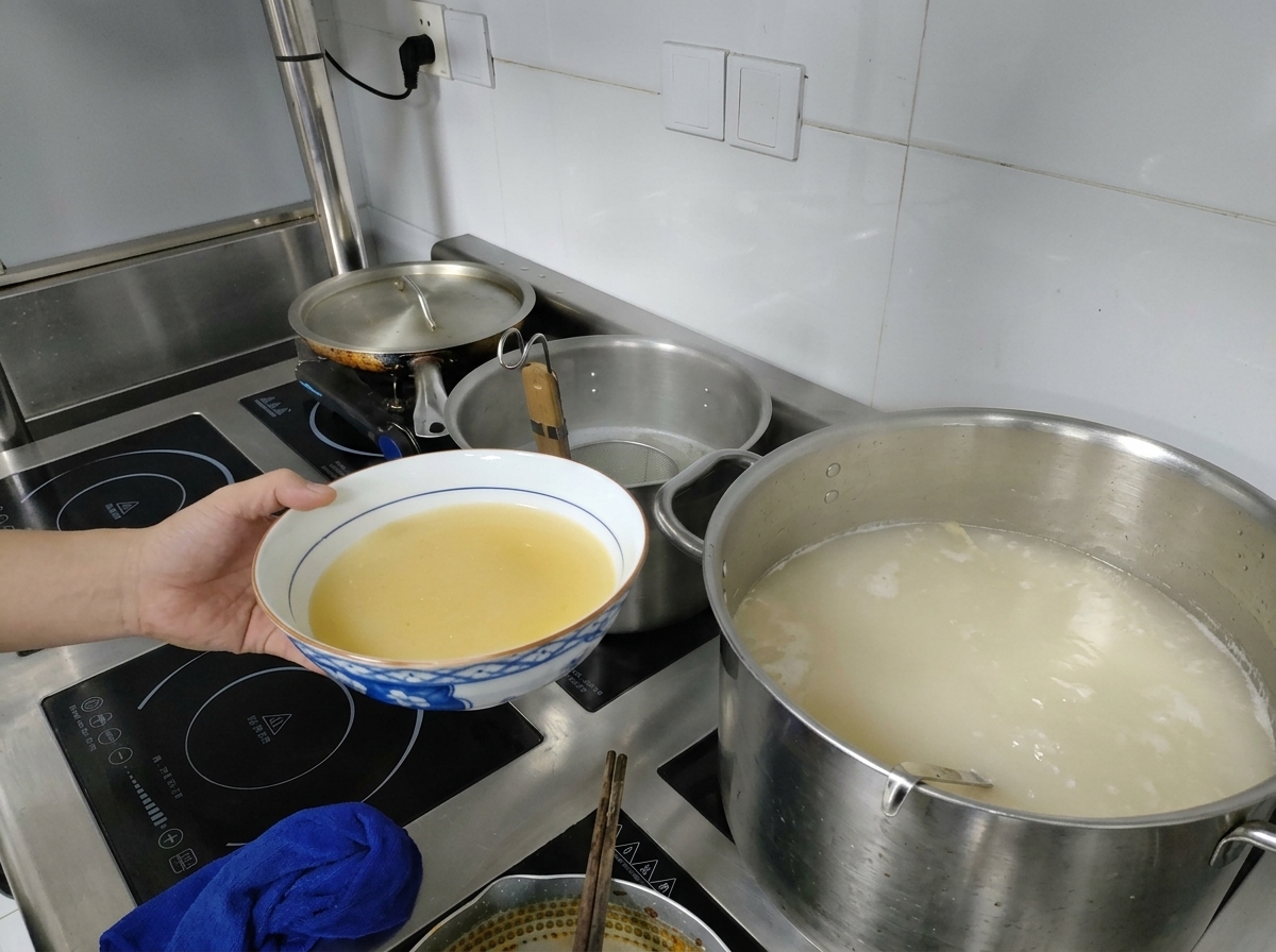 Ręka trzymająca miseczkę z żółtą pastą miso obok dużego garnka z gotującym się mlecznobiałym bulionem.
