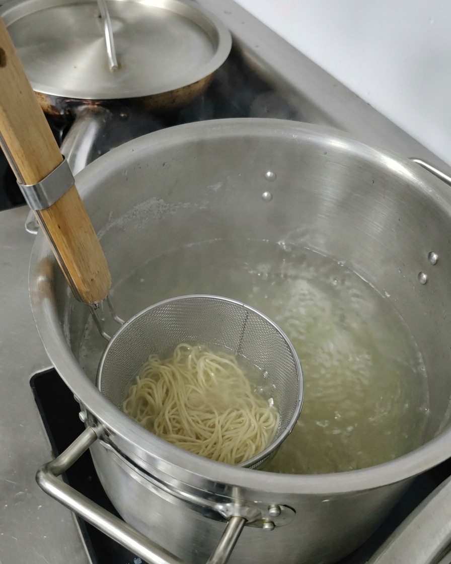 Wyjmowanie metalowego sitka z ugotowanym makaronem ramen z garnka z gorącą wodą.