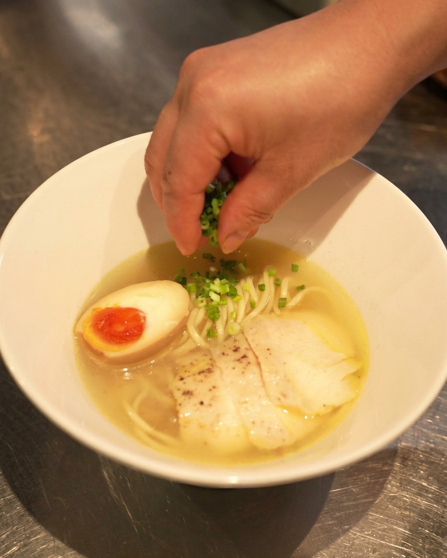 Dłoń posypująca gotowy ramen posiekaną zieloną cebulką.