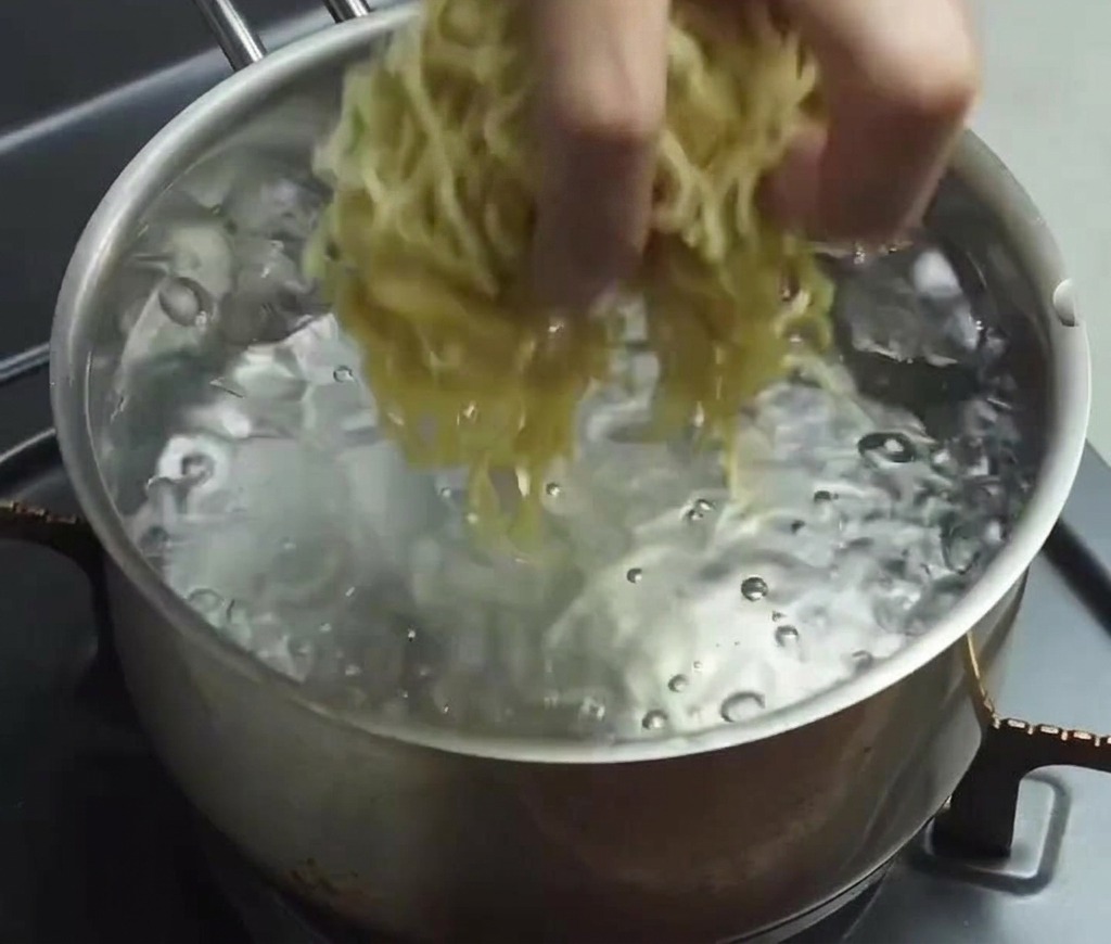 Dłoń wrzucająca porcję cienkiego, falowanego makaronu ramen do garnka z mocno wrzącą wodą.
