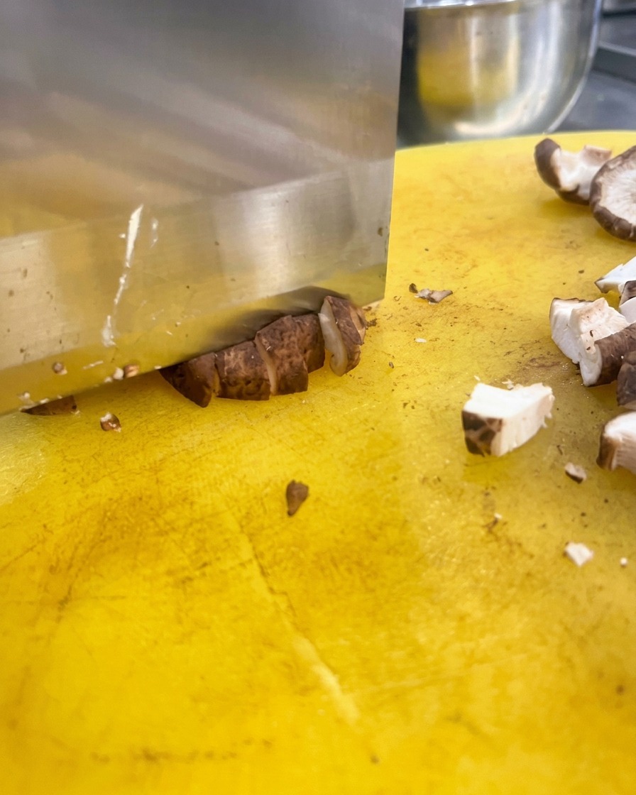Duży nóż kuchenny krojący kapelusze brązowych grzybów shiitake w drobną kostkę na jasnej żółtej desce do krojenia.