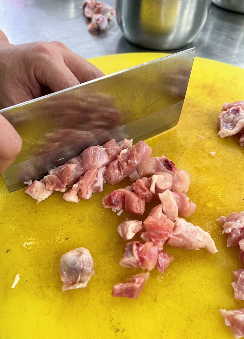 Osoba używająca dużego metalowego tasaka do krojenia surowego mięsa z kurczaka na żółtej plastikowej desce.