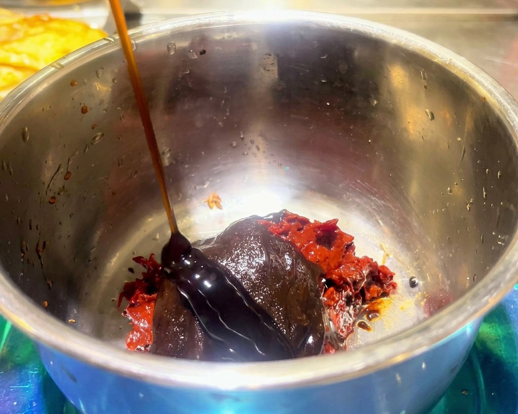 Gęsty ciemny sos wlewany na czerwoną pikantną pastę fasolową i przyprawę do hot pota w metalowej misce.