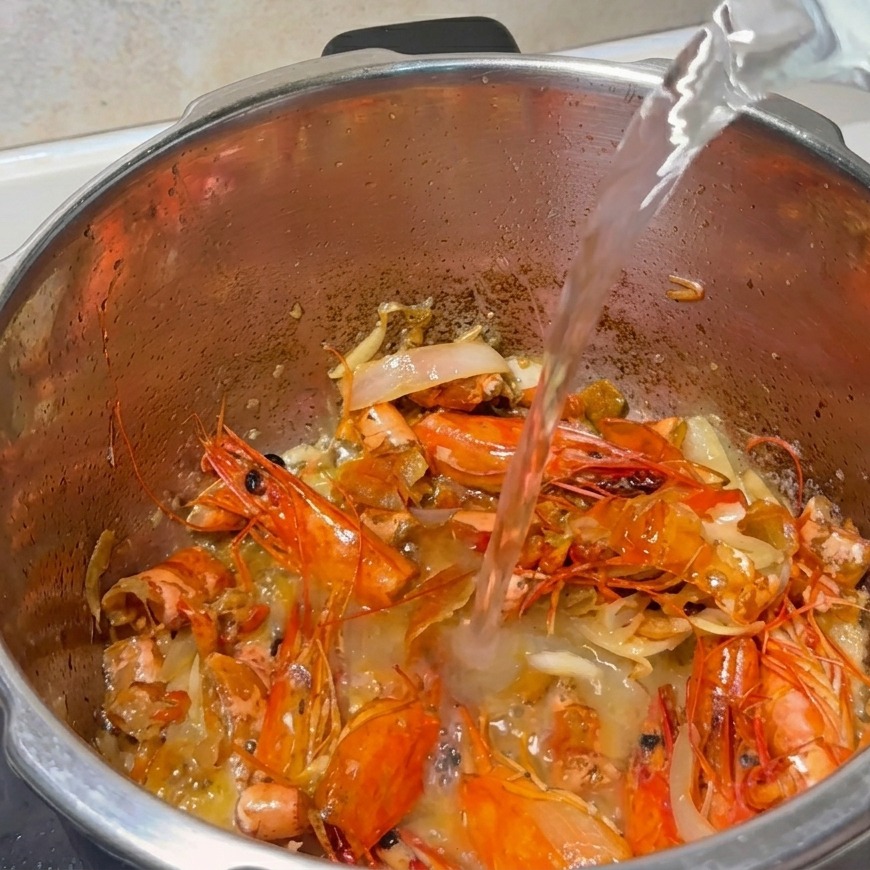 Strumień wody wlewany do garnka wypełnionego usmażonymi pancerzami krewetek i aromatycznymi warzywami.