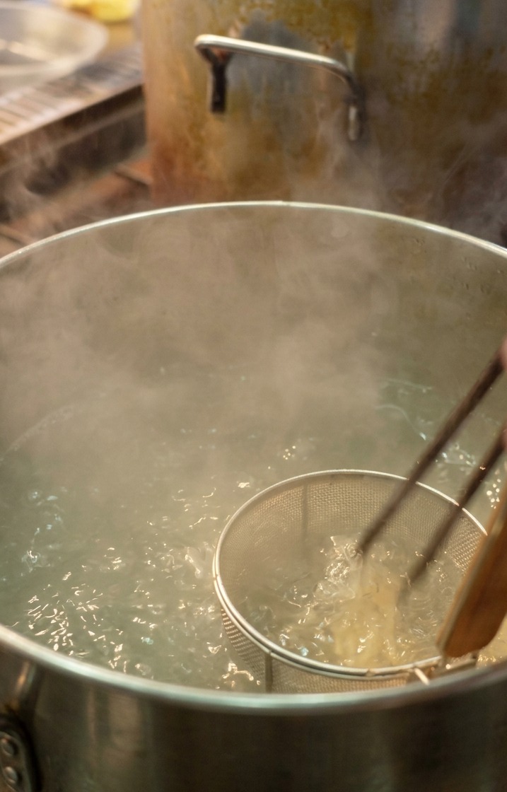 Szef kuchni używający sitka do makaronu, aby zanurzyć cienki biały makaron ramen w dużym garnku z wrzącą wodą.