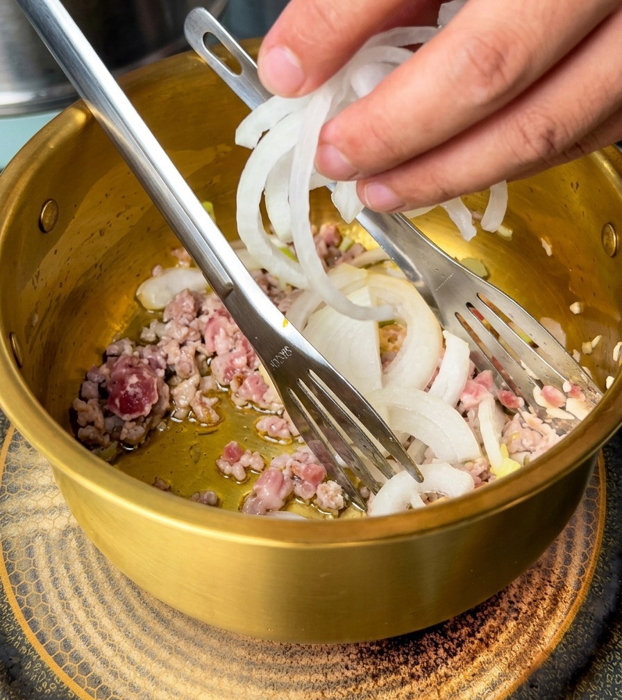 Dłoń dodająca pokrojoną białą cebulę do garnka z podsmażoną wieprzowiną i aromatami.