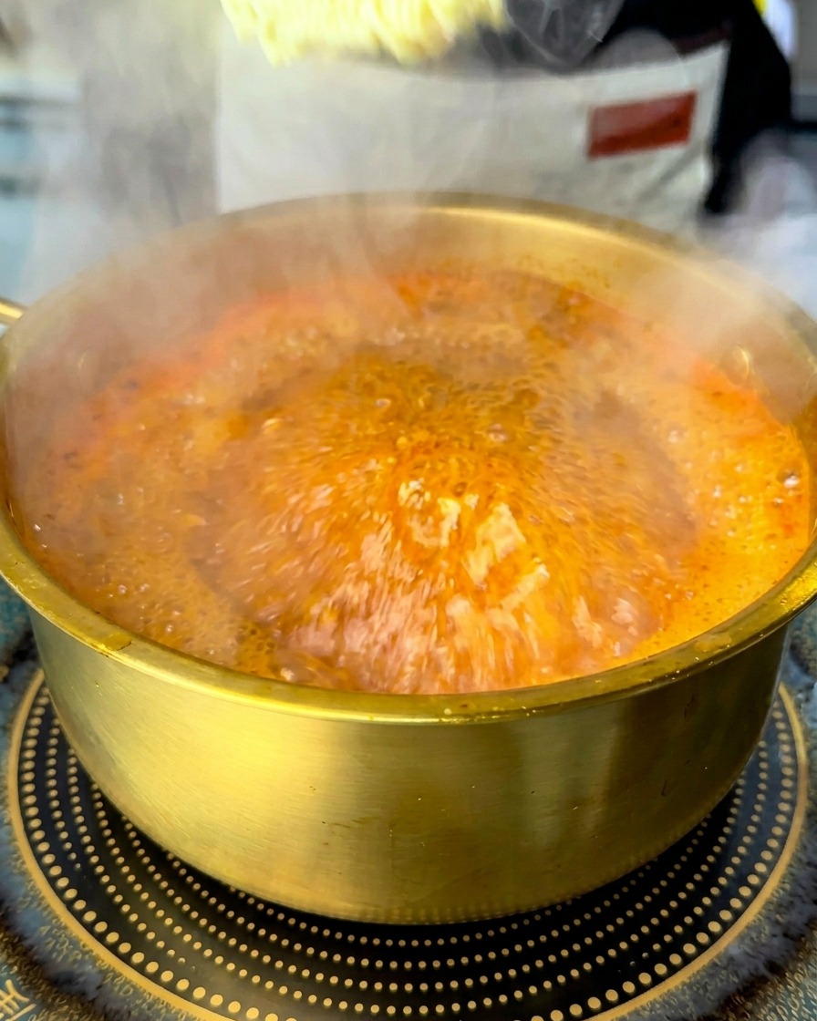 Blok suchego makaronu ramyun zanurzany w garnku z wrzącą czerwoną zupą.