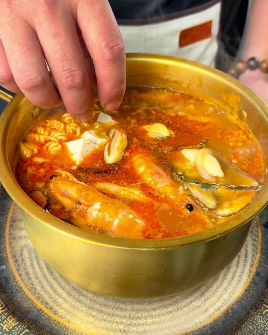 Dłoń wrzucająca kostki białego tofu i plasterki świeżego chili do gotującego się ramenu.