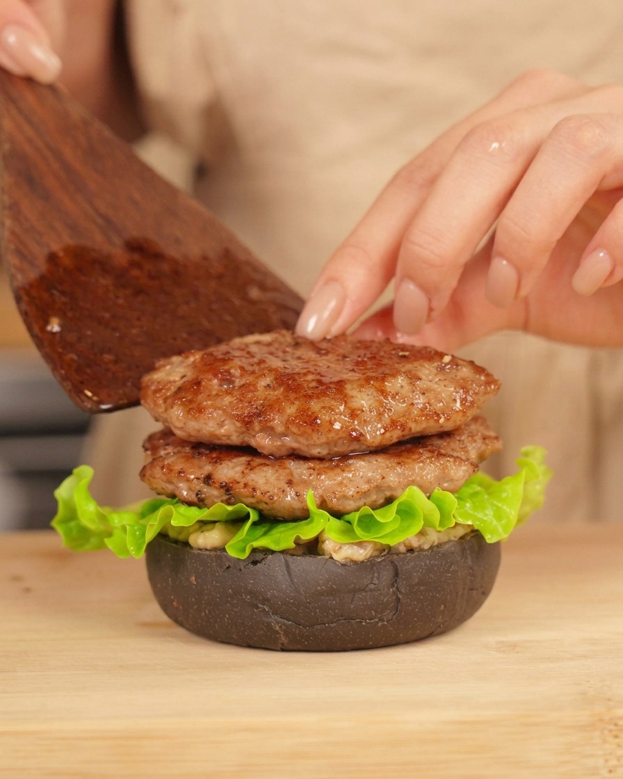 Osoba nakładająca łopatką drugi kotlet wołowy na częściowo złożonego burgera z sałatą.