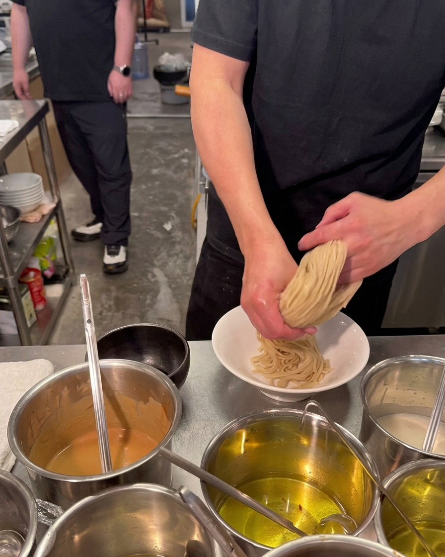 Kucharz oburącz składa i dociska porcję grubego, jasnego makaronu ramen w białej ceramicznej misce.