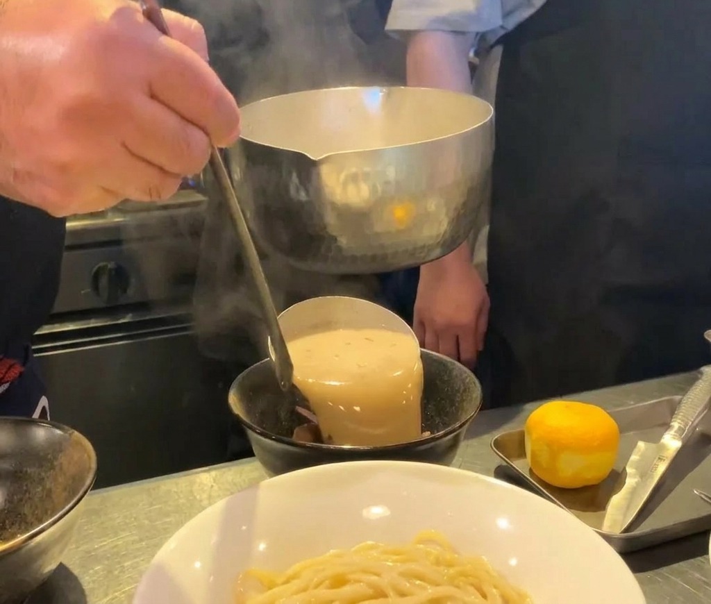 Gęsty, kremowy bulion ramen z owocami morza nalewany metalową chochlą do czarnej ceramicznej miseczki.