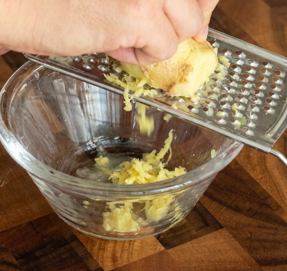 Dłoń trąca kawałek świeżego imbiru na tarce o drobnych oczkach, gładka pasta wpada do małej szklanej miseczki.