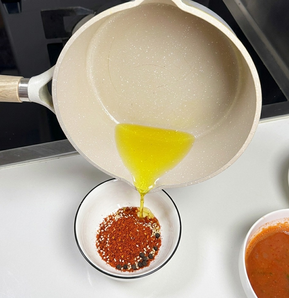 Jasny ceramiczny rondelek, z którego wylewany jest gorący, przezroczysty olej do małej białej miski wypełnionej mielonym chili i nasionami sezamu.