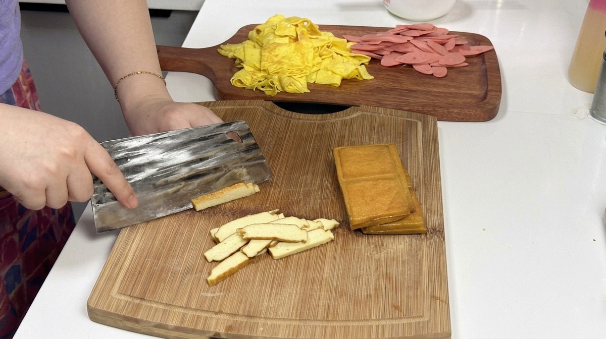 Metalowy tasak krojący stos złocistobrązowych bloczków wędzonego tofu na drewnianej desce, w tle widoczne przygotowane paski naleśników i szynki.