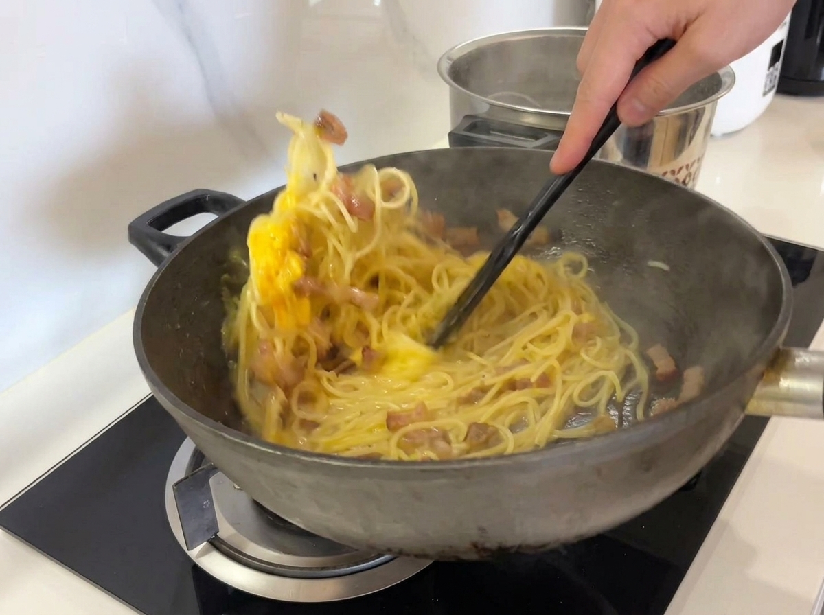 Energiczne mieszanie spaghetti, guanciale i masy jajecznej w woku przy użyciu czarnej łopatki.