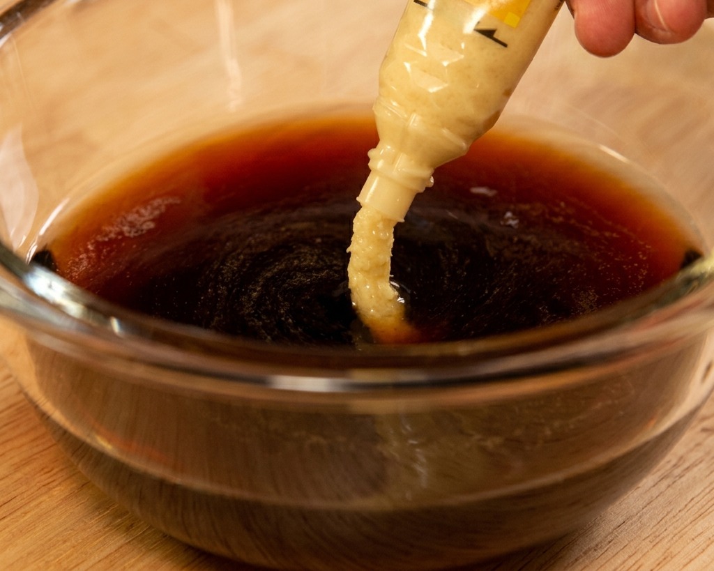 Pasta z tartego imbiru wyciskana z tubki do ciemnego sosu na bazie soi w szklanej misce.