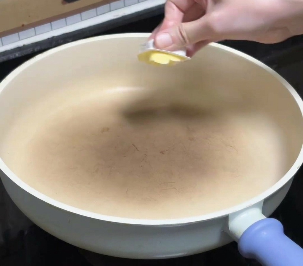 Dłoń kładąca kostkę żółtego masła na czystą, białą ceramiczną patelnię stojącą na kuchence.